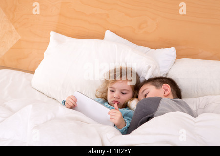 Müde, kleinen Bruder und Schwester kuscheln zusammen in einem warmen, bequemen Bett eine Gutenachtgeschichte auf einem Tablet Computing lesen Stockfoto