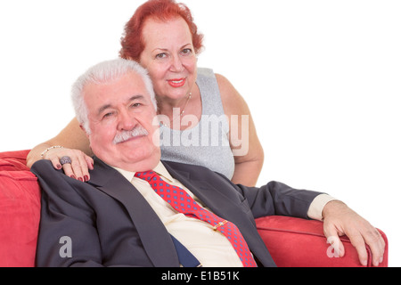 Älteres Ehepaar mit der Frau hinter ihrem Ehemann mit ihren Arm um seine Schulter sitzen gemeinsam auf einer roten Couch entspannen zu lieben Stockfoto