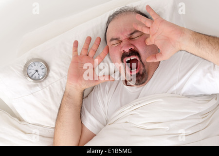Im mittleren Alter bärtiger Mann Gähnen laut Ruhe im Bett mit seinen Wecker auf dem Kissen neben ihm, als er versucht, nach dem Aufwachen Stockfoto