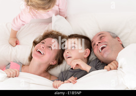 Verspieltes kleines Mädchen mit ihrer Familie im Bett klettern über den oberen Teil der Kissen, so dass sie mit Spaß und Vergnügen Lachen ein Stockfoto
