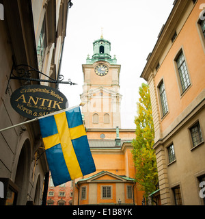 Eine schwedische Flagge wird vor einem Geschäft am Trångsund, als der Glockenturm der Storkyrkan (Stockholm Kathedrale), steigt hinter drapiert. Stockfoto