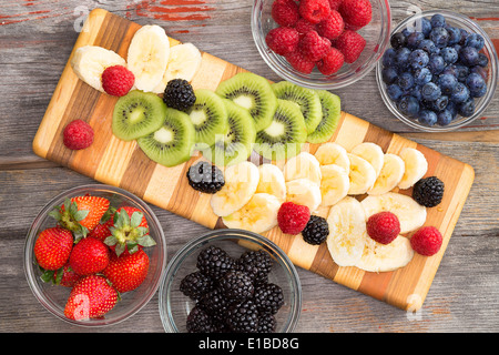 Vorbereitung einen gesunden Obstsalat mit gemischten Beeren in Einzelbehältnissen, in Scheiben geschnittenen Bananen und exotische Kiwi auf einem gestreiften woo Stockfoto