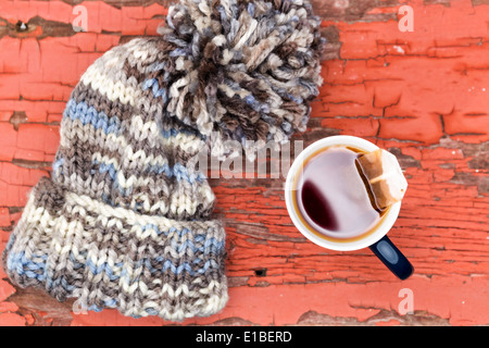 Blick von oben auf eine gemütliche gestrickte wollig Wintermütze mit einer großen Bommel neben einer Tasse frischen Tee auf einer Grunge Holzoberfläche Stockfoto