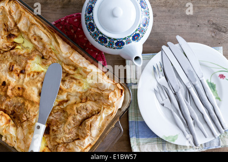 Draufsicht auf eine Auflaufform mit einer unbeschnittenen frisch gebackenen türkischen Borek bereit zum Servieren mit einem Keramik Teekanne und Haufen von plat Stockfoto