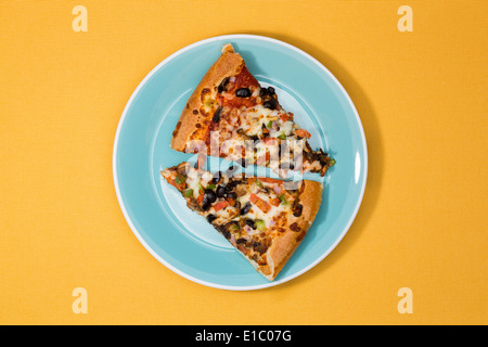 Draufsicht auf zwei Scheiben frisch gebackene herzhafte Pizza mit Käse, Tomaten, Oliven und Kräutern auf eine blaue Platte über einen gelben ba Stockfoto