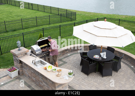 Erhöhte Ansicht eines Mannes Kochen von Fleisch auf ein Gas Grill steht in der Sonne auf einer gepflasterten Terrasse im Sommer Küche Prepaid Stockfoto