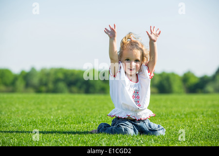 Lächelndes kleines Mädchen sitzt auf einem frischen grünen Rasen in einem Feld, Spaß mit ihren Händen in der Luft Stockfoto
