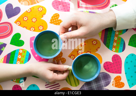 Womans und Mens Hände halten zwei Tassen Tee in der Hand auf einem Tisch mit Herz-Ornamenten Stockfoto