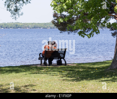 Junges Paar mit Arme umeinander auf Parkbank mit Blick auf den See. Stockfoto