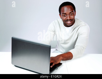 Lächelnder afrikanischen Mann am Tisch sitzen und mit Laptop auf grauem Hintergrund Stockfoto