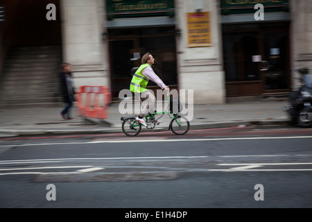 Mann trägt eine gelbe Jacke hi-viz Fahrten seine Brompton zusammenklappbar (Faltung) Fahrrad, mit einem vorderen Carrybag. Stockfoto