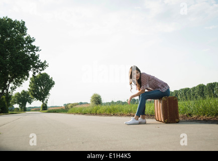 Junge Frau sitzt auf Koffer auf Landstraße Stockfoto