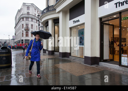 Eine Dame trägt Kopfhörer geht mit einem Regenschirm auf eine nasse Fahrbahn in Londons Strang. Stockfoto