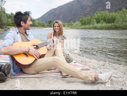 Freunde sitzen neben Fluss, Gitarre spielen Stockfoto