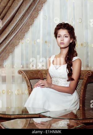 Weiblichkeit. Braun Haare Frau Braut im Hochzeitskleid sitzen. Klassische romantische Interieur Stockfoto