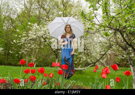 schöne blonde Mädchen mit blauen Kleid posiert im Park Blume Baum mit Regenschirm Stockfoto