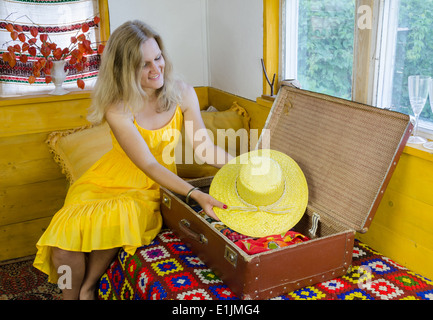 lächelnde Frau setzen schöne gelbe Sommerhut in alten Vintage Koffer im Village-Zimmer Stockfoto