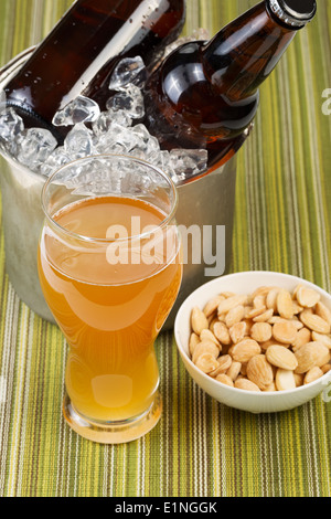 Closeup vertikale Foto von einem goldenen Bier in Longdrinkglas mit gesalzenen Nüssen in Schälchen mit Flaschenbier in Eimer mit Eis Stockfoto