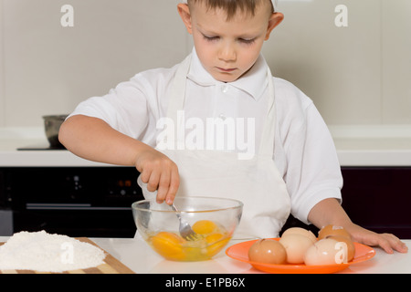 Kleiner Junge Eiern in einer Schüssel mischen, wie er in der Küche in seiner Schürze und weißer Haube steht, einen Kuchen zu backen Stockfoto