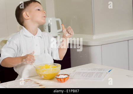 Kleiner Junge in der Küche aufgeben einen Daumen nach der Genehmigung steht er an der Theke mit seinem mischenden Schüssel und Zutaten Backen Stockfoto