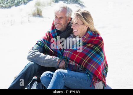Glückliches Paar eingehüllt in Decke am Strand sitzen Stockfoto