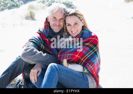 Glückliches Paar eingehüllt in Decke am Strand sitzen Stockfoto