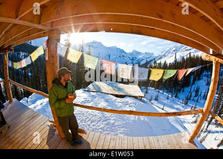 Ein Mann genießen Sie Sonnenaufgang und einen Kaffee im Backcountry-Ski-Hütte. Eisfall Lodge, Golden, BC Stockfoto