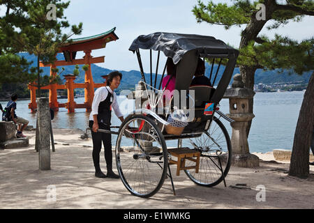 Läufer im Gespräch mit Touristen sitzen in seiner Rikscha auf Insel Miyajima Japan lächelnd. Im Hintergrund ist das Tor oder torii Stockfoto