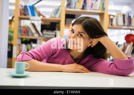 Porträt der jungen Frau mit Kaffee am Schreibtisch in Bibliothek Stockfoto