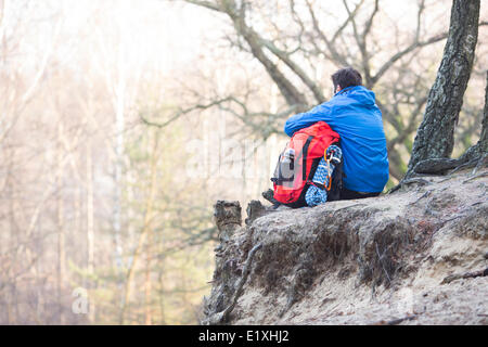 Rückansicht des Wanderer mit Rucksack sitzt am Rand der Klippe im Wald Stockfoto