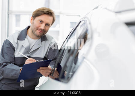Porträt von zuversichtlich Kfz Mechaniker Holding Zwischenablage beim stützte sich auf Autofenster in Werkstatt Stockfoto