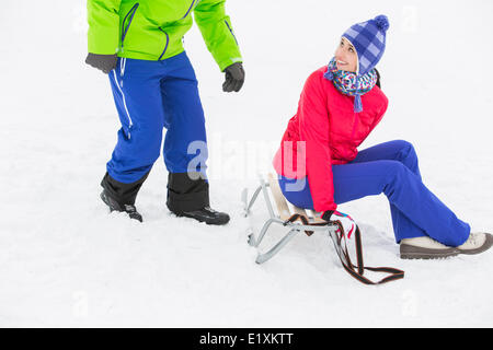 Glückliche junge Frau auf Schlitten sitzend und Blick auf den Mann im Schnee Stockfoto