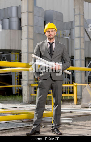 Porträt von zuversichtlich junger Geschäftsmann hält aufgerollt Blaupausen außerhalb Industrie Stockfoto