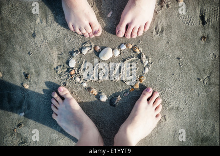 Füße von Liebespaar und ein Herz gemacht von Muscheln im Sand am Strand Stockfoto