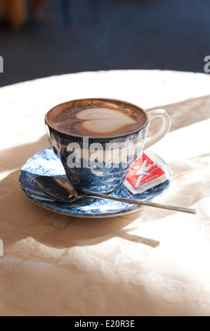 Einen Cappuccino in einer blau-weiß dekorierte Tasse mit einem Teelöffel und einem Beutel Zucker auf einem Tisch mit einem Leinentuch. Stockfoto