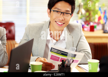 Glücklich asiatischen Geschäftsmann in Gläser am Tisch sitzen und halten Dokument im Büro Stockfoto