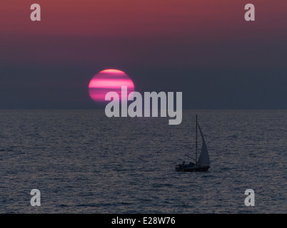 Eine Silhouette eines kleinen Segelboots in der Dämmerung mit einem großen roten und rosa gestreifte Sonne im Hintergrund Stockfoto