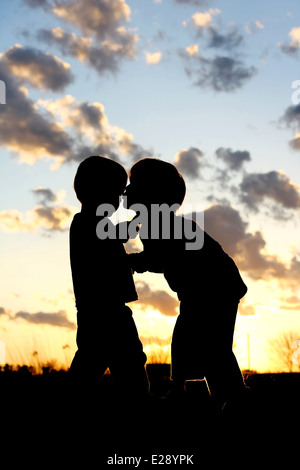 eine Silhouette eines jungen Kindes küssen seinen kleinen Bruder auf die Wange außerhalb bei Sonnenuntergang Stockfoto