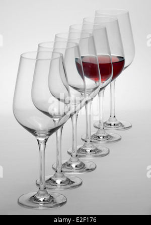 einige Gläser Wein stehen hintereinander in einer Reihe in hellem Grau zurück, eine teilweise mit Rotwein gefüllt Stockfoto