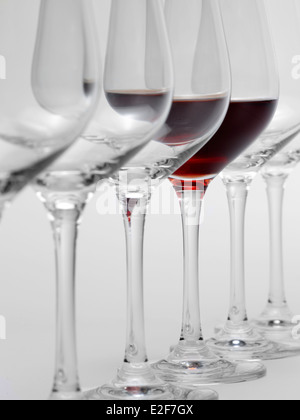 einige Gläser Wein stehen hintereinander in einer Reihe in hellem Grau zurück, eine teilweise mit Rotwein gefüllt Stockfoto