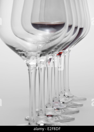 einige Gläser Wein stehen hintereinander in einer Reihe in hellem Grau zurück, eine teilweise mit Rotwein gefüllt Stockfoto