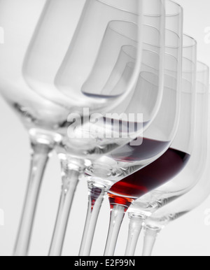 einige Gläser Wein stehen hintereinander in einer Reihe in hellem Grau zurück, eine teilweise mit Rotwein gefüllt Stockfoto