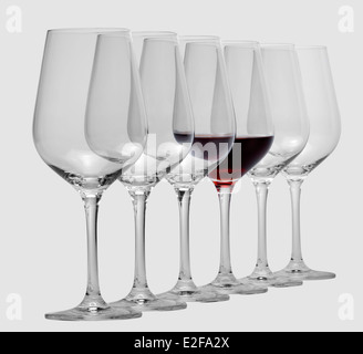einige Gläser Wein stehen hintereinander in einer Reihe in hellem Grau zurück, eine teilweise mit Rotwein gefüllt Stockfoto