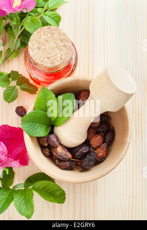 ätherisches Öl in Glasflasche, getrockneten Hagebutten Beeren in hölzernen Mörser und Hagebutte Blumen auf Tisch, Ansicht von oben Stockfoto