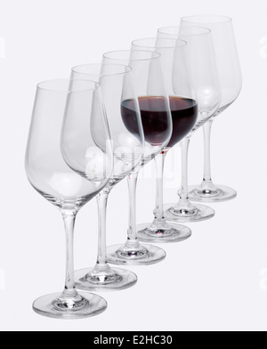 einige Gläser Wein stehen hintereinander in einer Reihe in hellem Grau zurück, eine teilweise mit Rotwein gefüllt Stockfoto