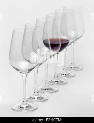 einige Gläser Wein stehen hintereinander in einer Reihe in hellem Grau zurück, eine teilweise mit Rotwein gefüllt Stockfoto