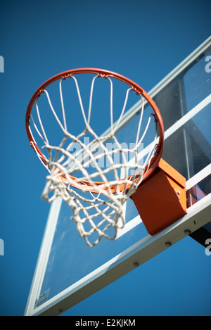 Basketballkorb Orange Netz Blauer Himmel | Ein Basketballkorb und ein herabhängendes Netz werden aus einem steilen Tiefwinkel gesehen, der orangefarbene Ring und der Pfahlhalter stehen im Kontrast zu einem hellblauen Himmel und der zerkratzten Acryloberfläche des Rückboards. Die sichtbare Befestigungsklammer und die Hardware heben die praktische Konstruktion hervor, die hinter der Befestigung eines regulären Korbs an seiner Stützstruktur steckt. Stockfoto
