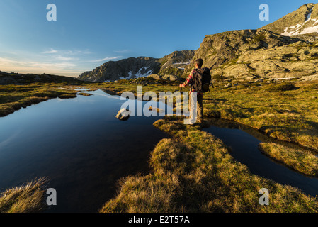 Männliche Wanderer mit Rucksack und Stick Rast und Sonnenaufgang in der Nähe von See und Bach in den italienischen Alpen zu beobachten. Stockfoto