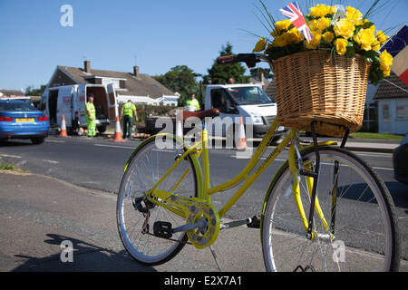 Frauen mit Blumen dekoriert gelb Fahrrad in York, Yorkshire, Großbritannien. 21. Juni 2014. Wartung Mannschaft Durchführung Samstag Straße Reparaturen auf der Tour de France Route. Insgesamt £ 4 Mio. wurde auf acht Yorkshire Räte zugeteilt wurden Straßen der Region zu erhalten, repariert und die Route bereit für die Tour de France im Juli. Das Geld wird ausgegeben, um sicherzustellen, dass die 389 Kilometer Asphalt für die Tour 2014 Grand abzuweichen, wird Schlagloch frei sein. Die Route wird nun mit gelben Blumen, Körbe gesäumt, die Bunting und Fahrräder. Stockfoto