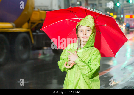 Junges Mädchen im Regen trägt einen grünen Regenmantel und hält einen roten Regenschirm auf Bürgersteig Stockfoto